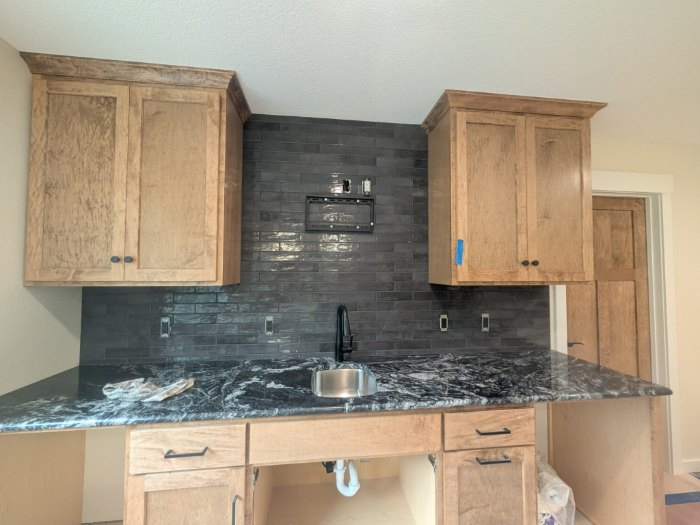 Dark charcoal glossy brick style tile backsplash running up the wall between wood cabinets, above a black and white granite counter