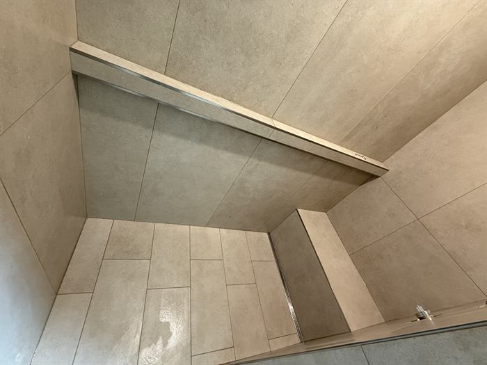 Upward view of a large format greige tile shower with a metal trimmed ledge and linear drain